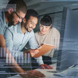 An IT professional and two smiling employees standing by a computer in a dimly lit office, showing the concept of an IT managed service provider (MSP)