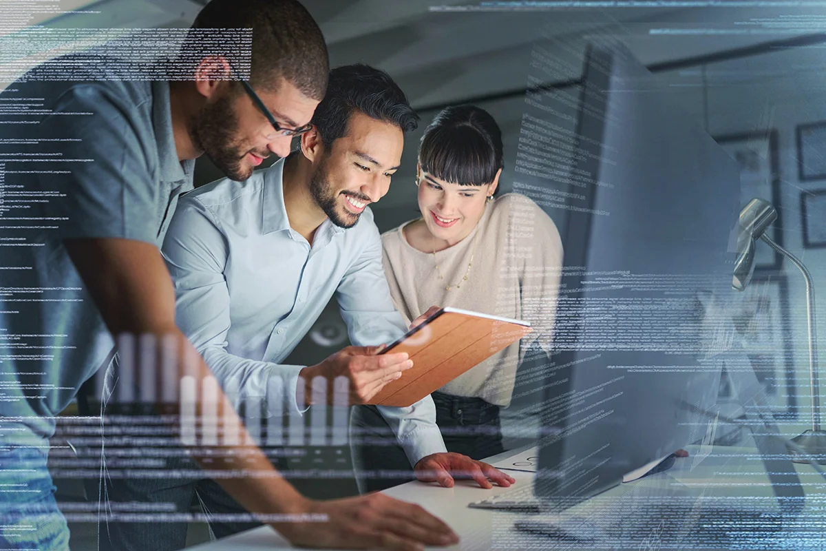 An IT professional and two smiling employees standing by a computer in a dimly lit office, showing the concept of an IT managed service provider (MSP)